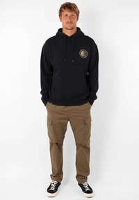 Sweat à capuche marine avec une poche frontale et un détail logo, associé à un pantalon cargo beige clair avec des poches latérales et des baskets noires.
