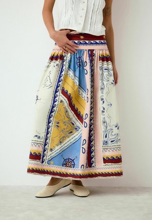Nő színes, mintás maxi szoknyában, tengeri és paisley mintákkal, fehér ujjatlan felsővel és bézs balerinacipővel párosítva.