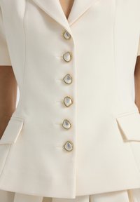 Blazer sartorial color crema con sei bottoni in perla di forma irregolare incorniciati in oro e tasche a patta frontali.