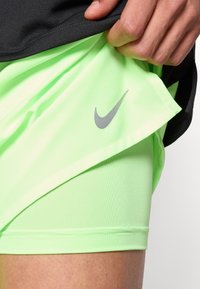 Φωσφοριζέ πράσινο αθλητικό σορτς με λεία υφή και γκρι λογότυπο Nike, κάτω από μαύρο πουκάμισο. Έμφαση στα αθλητικά σχεδιαστικά στ οιχεία.