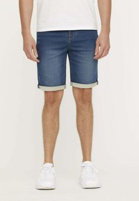 Shorts en denim bleu foncé avec ourlets repliés, présentant un design classique à cinq poches et une texture lisse, associés à des baskets blanches.