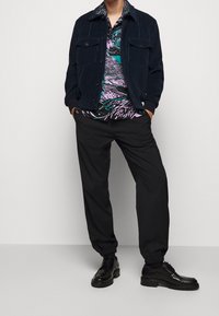 Veste en velours côtelé bleu foncé, chemise à motifs avec un design violet et vert, pantalon noir fuselé et chaussures noires à lacets.