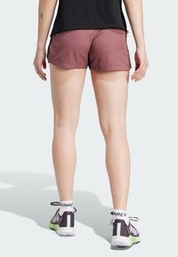 Burgunderfärgad sportshorts med en slät textur, kombinerat med mörka sneakers som har vita och gröna accenter. Synlig branding på strumporna.