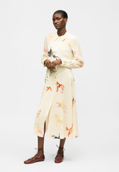 Γυναίκα φοράει κρεμ midi φόρεμα με κουμπιά και πολύχρωμα σχέδια αλόγων, συνδυασμένο με καφέ δερμάτινα παπούτσια με κορδόνια, στέκεται μπροστά σε λευκό φόντο.