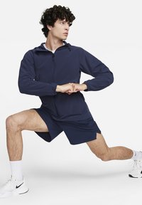 Donkerblauwe Nike zip-up jas met capuchon, gecombineerd met bijpassende shorts. Kenmerkt zich door een slim fit, een soepele textuur en een subtiele logo op de borst.