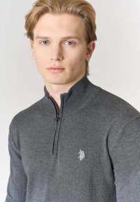 Maglione grigio con zip in morbido materiale. Presenta un collo alto e un piccolo logo ricamato sul petto a sinistra.