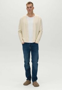 Svetlo béžový cardigan, biele tričko s okrúhlym výstrihom, modré riflové nohavice a hnedé slip-on topánky. Cardigan má zapínanie na gombíky a rebrované manžety.