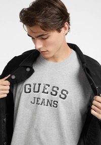 Pilkas medvilnės marškinėliai su tamsiu, nusidėvėjusiu „GUESS JEANS“ tekstu, dėvimi po juodu džinsiniu švarku su užsegamu apykakle ir tekstūruota medžiaga.