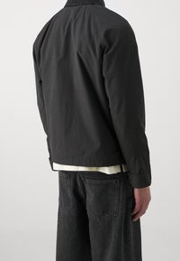 Person iført en sort jakke over en hvid skjorte med løstsiddende mørkegrå jeans, vist fra ryggen mod en ensfarvet grå baggrund.
