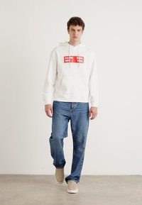 Hanorac alb cu text grafic roșu, cu șnururi la gât, purtat cu blugi albaștri și adidași de culoare deschisă. Design minimalist.