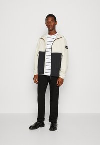 Lichtbeige en zwarte lichte jas met een capuchon, ritssluiting aan de voorkant en twee zijzakken. Draag het over een wit met zwart gestreept T-shirt. Zwarte broek en schoenen.