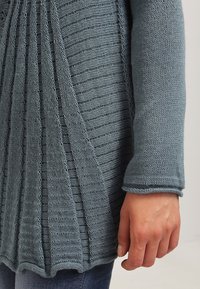 Ljusblå stickad tröja med vertikala ribbdetaljer, långa ärmar och en avslappnad passform. Tyget verkar mjukt och strukturerat.