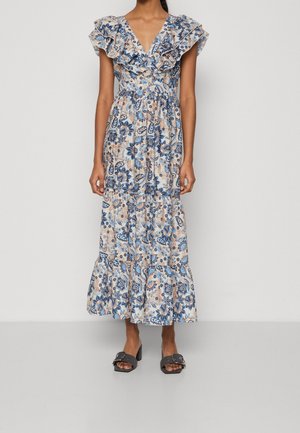 Femme portant une robe longue fleurie sans manches avec un décolleté en V à volants et une jupe à étages, assortie à des sandales noires à talons ouverts.