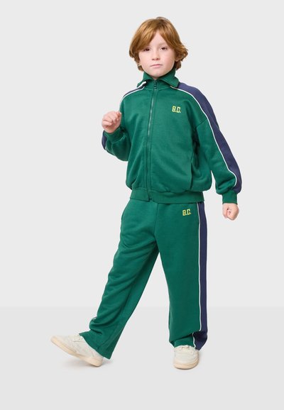 Zeleni trener s tamnoplavim detaljima, koji se sastoji od jakne na patentni zatvarač i hlača s elastičnim pojasom. Uključuje bijele pruge i detalj s logom. Bijele tenisice.