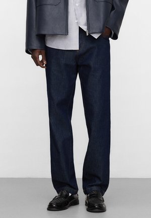 Mann trägt dunkelblaue Jeans mit geradem Bein, schwarze Lederloafer, ein gestreiftes Hemd und eine marineblaue Jacke, die Hände teilweise in den Taschen.