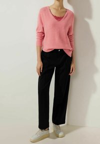 Rosa v-ringad tröja i texturerad stickning, lager på lager med en topp med spetsdetaljer. Svarta byxor med vida ben och ljusa sneakers kompletterar outfiten.