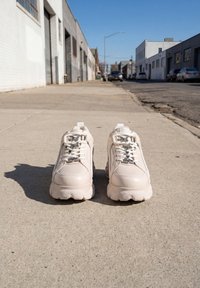 Hvide chunky sneakers med butterfly og hjerte snørelås vedhæng placeret side om side på en solbeskinnet byfortov.