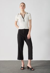 White ribbed short-sleeve top with a black zip and collar, paired with black cropped trousers and black heeled sandals with a twisted design.
