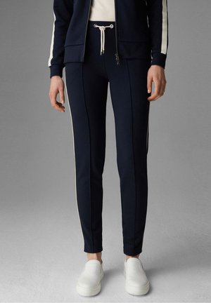 Persoon draagt marineblauwe slim-fit broek met witte zijstrepen, een bijpassend marineblauw jasje, witte top en witte instapschoenen tegen een grijze achtergrond.