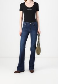 Žena v černém topu "GUESS JEANS", tmavě modrých rozšířených džínách, hnědých špičatých botách, držící olivově zelenou kabelku na bílém pozadí.