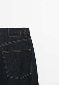 Poche arrière et passants de ceinture d'un jean en denim bleu foncé avec surpiqûres orange contrastantes sur fond blanc.