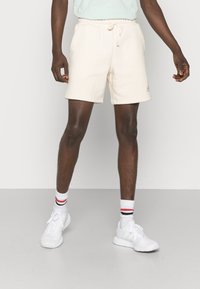 Beżowe bawełniane szorty z regulowanym sznurkiem w pasie, bocznymi kieszeniami i detalem z logo. Kompletnie zestawione z białymi trampkami i podkolanówkami w czerwone i czarne paski.