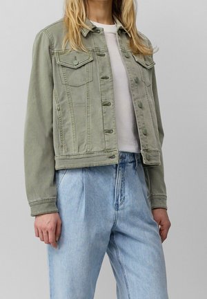 Femme portant une veste en jean vert clair boutonnée sur un t-shirt blanc et un jean bleu clair ample sur un fond uni.