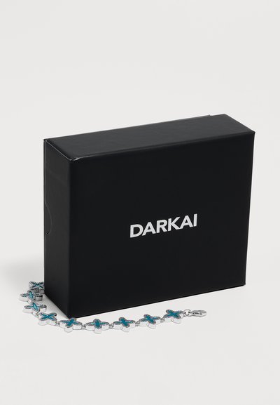 Boîte rectangulaire noire avec le nom de la marque Darkai en blanc, accompagnée d'un bracelet en argent avec des maillons bleus en forme de croix.