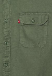 Camicetta button-up verde scuro realizzata in tessuto testurizzato, con una tasca sul petto con pattina, due bottoni e una piccola etichetta con logo.