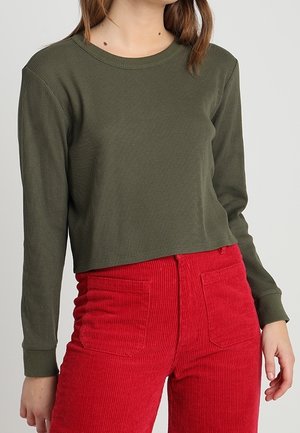 Kobieta ubrana w oliwkowozielony, prążkowany crop top z długim rękawem oraz czerwone, sztruksowe spodnie z wysokim stanem i przednimi kieszeniami.