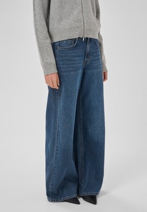 Personne portant un pull zippé gris clair et un jean bleu à jambes larges, debout bras détendus, chaussures noires pointues à peine visibles.