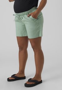 Personne portant un short à cordon de serrage vert clair avec des poches et des tongs à plateforme noires, se tenant contre un fond uni.