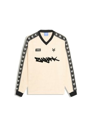 Chemise beige à manches longues avec col en V noir, poignets noirs, rayures noires avec logos blancs sur les manches et texte graphique noir à l'avant.