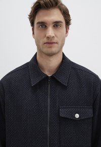 Marineblaues strukturiertes Jacket mit Reißverschluss, spitzem Kragen und Brusttasche. Verfügt über einen silbernen Knopf und ein dezentes 3D-Muster.