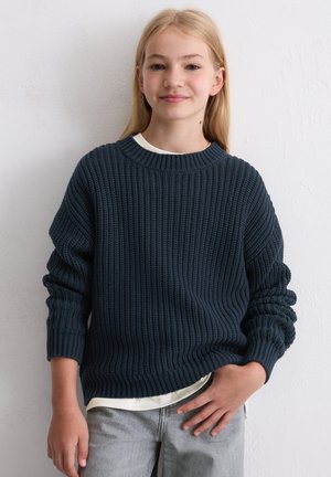 Blondes Mädchen trägt einen dunkelblauen grob gestrickten Pullover, darunter ein weißes Hemd und helle graue Jeans, steht vor einer schlichten weißen Wand.