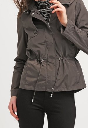 Frau trägt eine dunkelgraue Jacke mit Reißverschluss und Kordelzug in der Taille über einem schwarz-weiß gestreiften Rollkragenpullover und schwarzen Hosen.