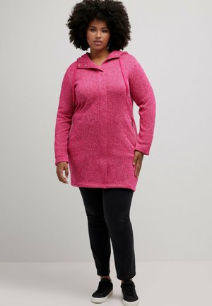 Veste en polaire rose avec fermeture éclair à l'avant, capuche ajustable, deux poches latérales et tissu textured. Associée à un pantalon noir et des baskets.
