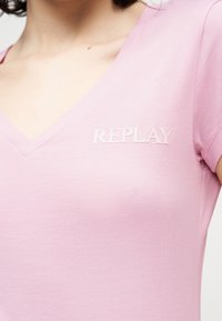 Κοντινό πλάνο ενός ατόμου που φοράει ένα ροζ ανοιχτό T-shirt με V λαιμόκοψη και την επιγραφή "REPLAY" σε λευκό χρώμα στο πάνω αριστερό μέρος του στήθους.