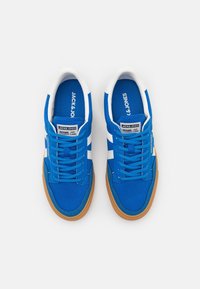 Blå canvas sneakers med vita ränder, rund tå och brun gummisula. Har skosnören och logotyp på tungan.