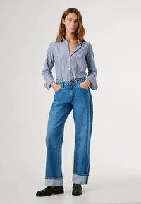 Pruhovaná košile na knoflíky v modré a bílé, kombinovaná s vysokým pasem modrých džín s ohrnutým lemem. Černé boty doplňují outfit.