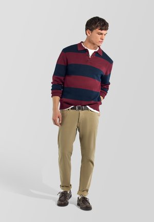 Joven con una camisa de manga larga a rayas burdeos y azul marino, pantalones beige y zapatos marrón oscuro, de pie frente a un fondo gris claro.