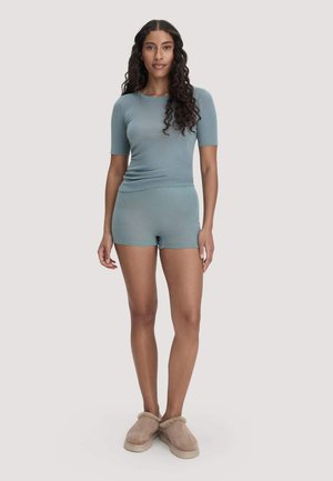 Femme aux longs cheveux bouclés portant un haut bleu ajusté à manches courtes, un short assorti et des chaussons beige à enfiler, debout sur un fond clair uni.