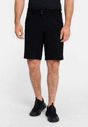 Mann trägt schwarze legere Shorts, schwarze Slip-On-Sneaker und ein schwarzes Hemd, steht vor einem schlichten hellen Hintergrund.