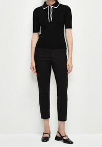 Polo en maille côtelée noire avec col blanc et liens, associé à un pantalon noir ajusté et des chaussures Mary Jane noires. Texture lisse, coupe ajustée.