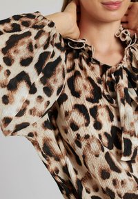 Blusa de estampado de leopardo con mangas abullonadas, textura plisada y cuello atado. Los colores incluyen beige, negro y acentos marrones.