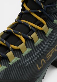 Melna pārgājienu kurpe ar teksturētu virsmu, kas ir apvīta ar dzeltenām un tumši zilām saitēm, zaļiem akcentiem un "LA SPORT" zīmolu zeltītā krāsā.