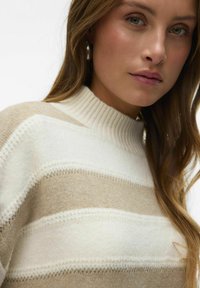 Pull rayé beige et crème avec un col haut à côtes, présentant une texture douce et des détails subtils dans le motif de tricot.