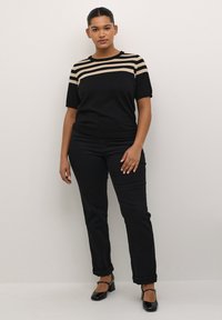 Chemise noire avec des rayures beiges aux épaules, associée à un pantalon noir fuselé. La mannequin porte des chaussures à talons blocs noires. Design simple et ajusté.