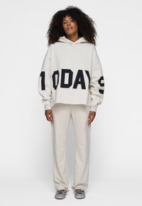 Sudadera corta gris claro con texto negro "1 HOY" en el frente y en la manga izquierda, combinada con pantalones de chándal grises, creando un look de ropa de estar en casa cohesivo.