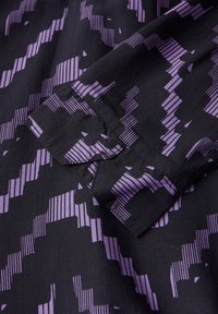 Tissu noir avec un motif géométrique violet, comportant des rayures verticales et en zigzag. Texture lisse avec une manche pliée visible.